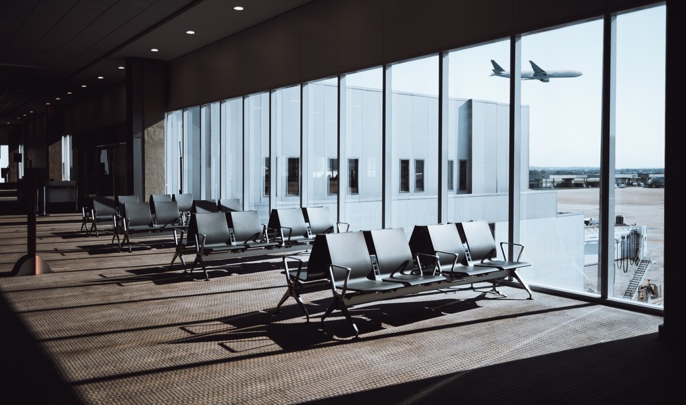 empty airport terminal seating area with rows of chairs facing large windows, through which a plane can be seen taking off